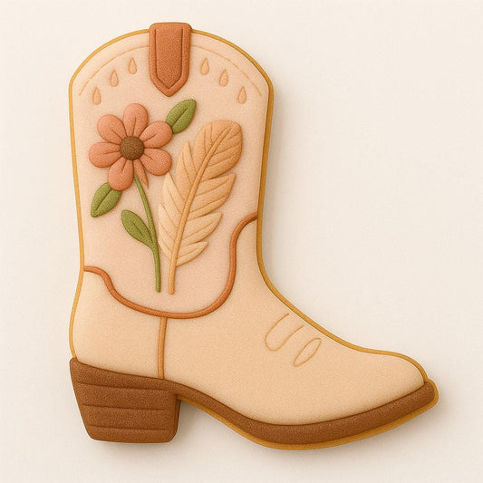 A tan western boot–shaped cookie decorated with soft peach icing, a brown sole and heel, and a floral pattern featuring a pink flower, green leaves, and a feather. The design combines rustic cowboy charm with a delicate boho aesthetic.
