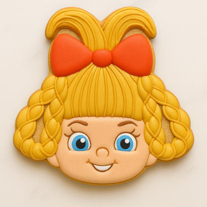 A cookie shaped like a smiling blonde girl with big blue eyes, braided hair, and a large red bow on top. The design captures a cheerful Whoville-style character, evoking festive holiday charm and classic Christmas nostalgia.