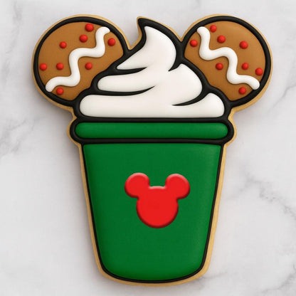 A cookie shaped like a green holiday latte cup topped with white whipped cream and two gingerbread mouse ears decorated with red and white icing. The front of the cup features a red mouse silhouette, creating a festive Disney-inspired Christmas cookie design.