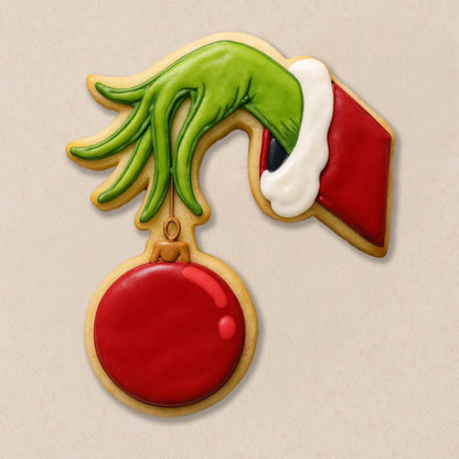 A festive cookie set inspired by The Grinch, including cookies shaped like the Grinch in a Santa hat, Max the dog with an antler, a Who girl with blonde braids and red bow, a crooked green Christmas tree with lights, and the Grinch’s green hand holding a red ornament.