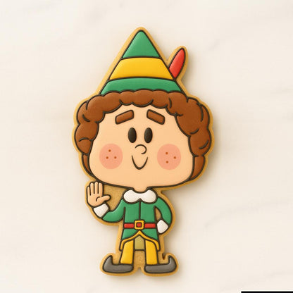 A cookie shaped like a smiling Christmas elf wearing a green outfit with yellow accents, a striped green-and-yellow hat with a red feather, rosy cheeks, and brown curly hair, standing with one hand raised in a friendly wave.