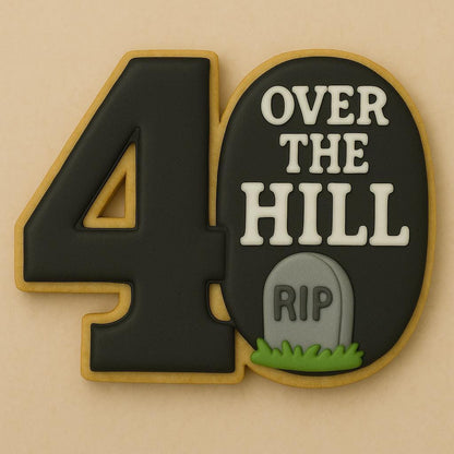 A sugar cookie shaped like the number 40, decorated with black icing and white letters reading “OVER THE HILL,” featuring a small gray tombstone with “RIP” written on it and a patch of green grass.