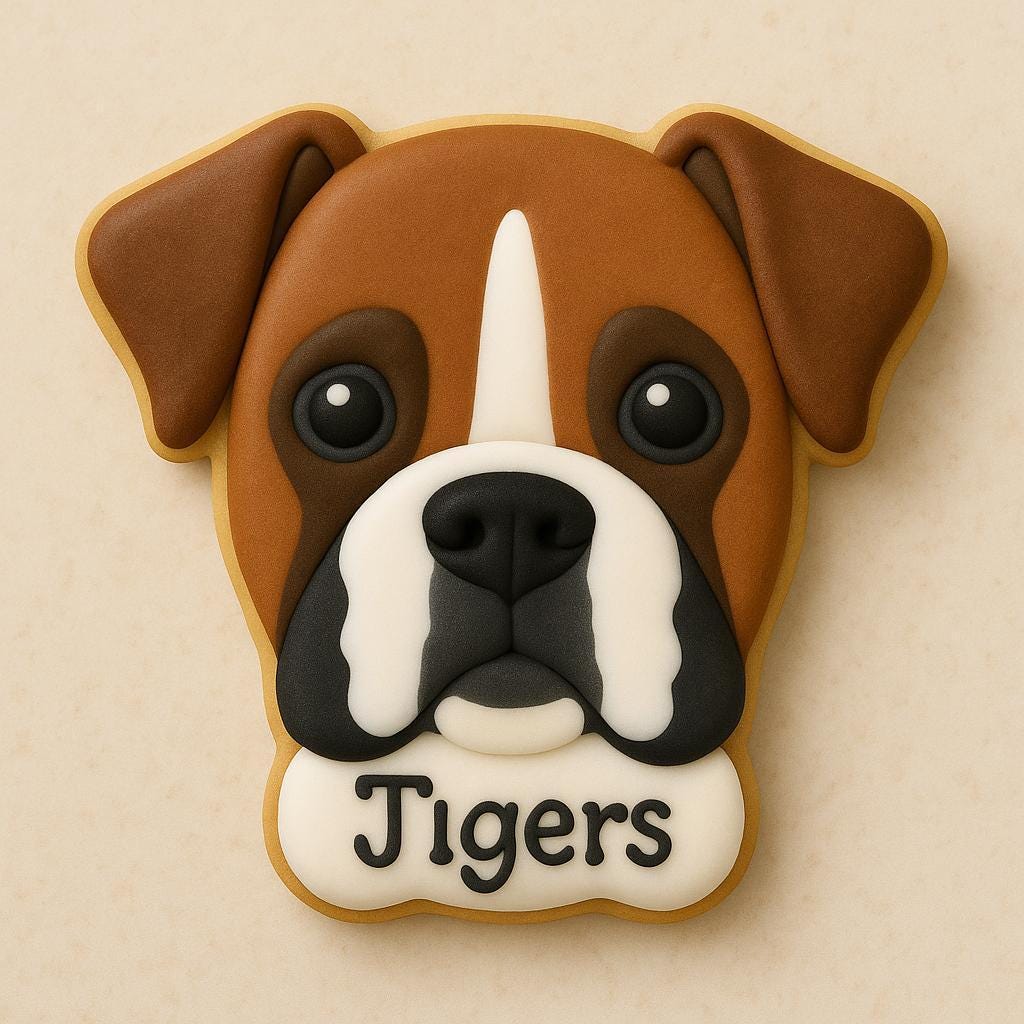 A sugar cookie shaped like a Boxer dog’s face, decorated with brown, white, and black icing details, large dark eyes, and the word “Tigers” piped in black icing at the bottom.