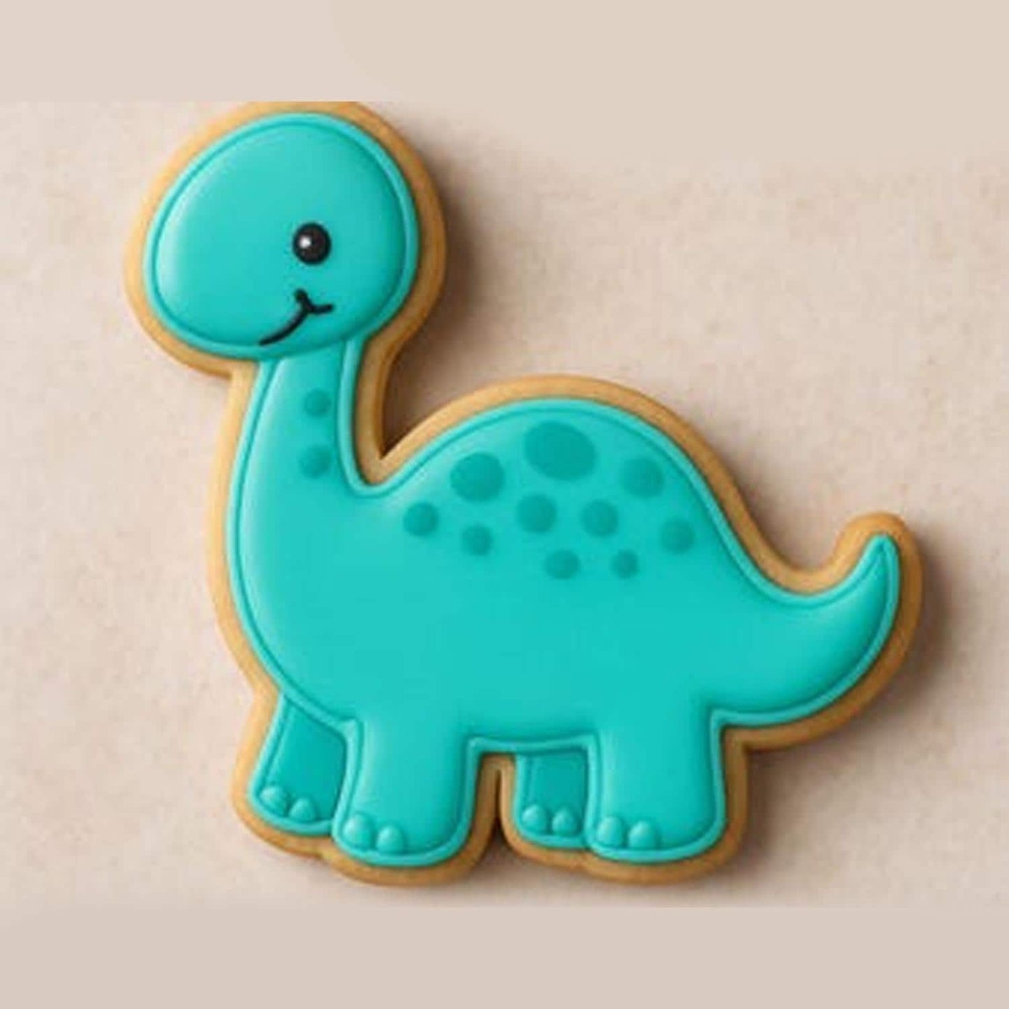 A sugar cookie shaped like a smiling turquoise Brontosaurus with darker blue spots and black eyes, set against a light beige background.