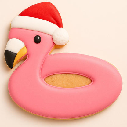 A pink flamingo-shaped cookie decorated with a red Santa hat and white fluffy trim. The flamingo has a black-and-yellow beak and a hollow center resembling a pool float. The design combines tropical and festive holiday themes, perfect for a warm-weather Christmas.