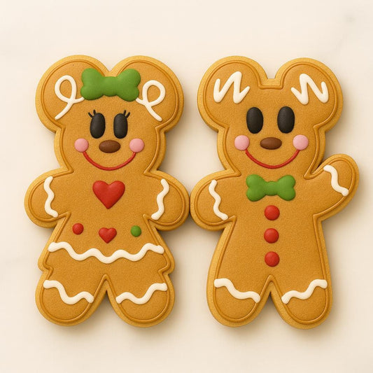 A matching set of two gingerbread cookies — one styled as a girl with a green bow, red heart, and icing skirt trim, and one styled as a boy with a green bow tie and red buttons. Both have smiling faces, pink cheeks, and white icing details, creating a festive and adorable holiday pair.