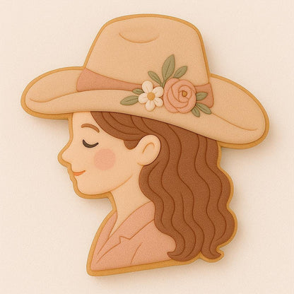 A cowgirl-shaped cookie decorated with a tan hat featuring floral accents, flowing brown hair, and soft blush tones. The design shows a side profile of a serene, smiling cowgirl with a classic western style and elegant boho details.