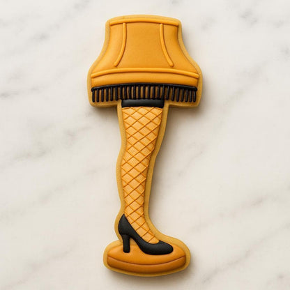 A cookie shaped like the famous leg lamp from A Christmas Story, featuring a golden lampshade with black fringe, fishnet-patterned stocking, and black high heel — a fun and nostalgic holiday cookie design.