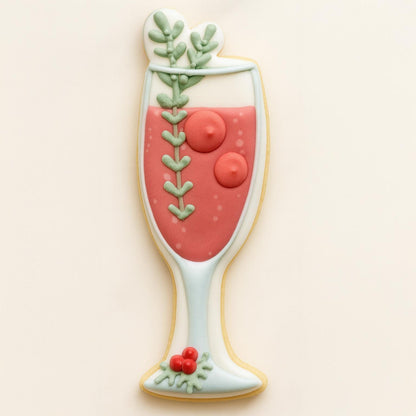 A cookie shaped like a champagne flute filled with a pink holiday cocktail, decorated with green leaves, red berries, and a holly accent — a festive and elegant Christmas cookie design.