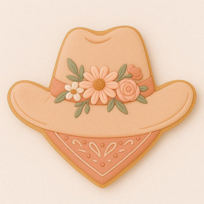 A sugar cookie shaped like a wide-brimmed cowboy hat with a floral crown of peach, cream, and sage flowers, resting above a paisley bandana in soft terracotta tones.