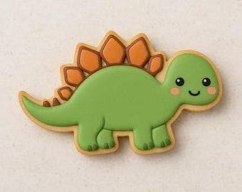 A sugar cookie shaped like a green stegosaurus with orange plates and a smiling face, decorated with pink cheeks, set on a neutral background.